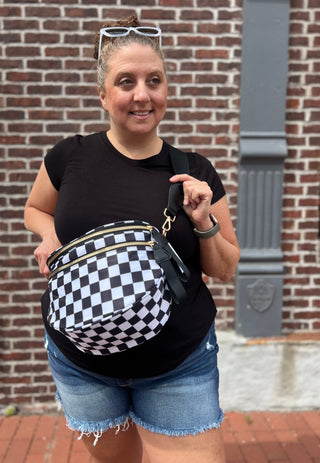 A woman wearing a black t-shirt and denim shorts models a black and white checkered fanny pack with gold-tone hardware, standing in front of a brick wall.