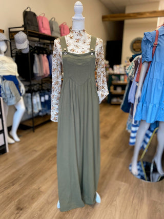 A woman's outfit displayed on a mannequin inside a clothing store. The outfit consists of an olive green overall-style maxi dress worn over a long-sleeve cream floral print shirt.