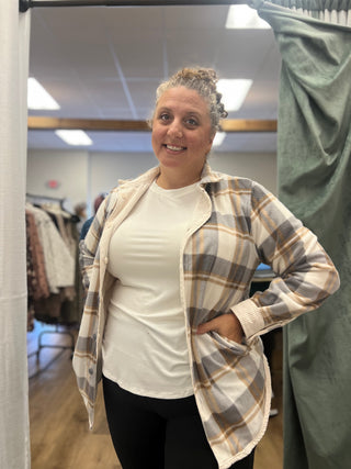 A woman modeling a plaid button-up overshirt with a white t-shirt underneath, standing in a fitting room or boutique.