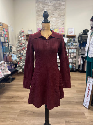 A ribbed knit long-sleeve collared dress in deep burgundy displayed on a mannequin inside a boutique. The dress features a buttoned collar and a flared skirt, set against a background of wooden floors and store fixtures.