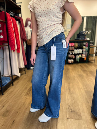 A person wearing blue wide-leg jeans and a light beige floral top stands in a clothing store, with the jeans still showing white retail tags. Clothing racks, mannequins, and a wood floor are visible in the background.