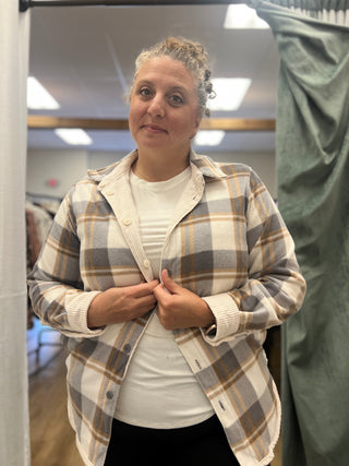 A woman wearing a beige, brown, and gray plaid button-up shirt over a white ribbed t-shirt, standing in a fitting room.