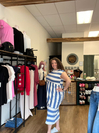 A woman wearing a blue and white striped dress takes a mirror selfie inside a clothing store. Clothing racks with various garments are visible in the foreground, and the store interior with tiled walls and wooden columns is in the background.