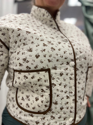 A close-up of a person wearing a quilted jacket with a brown floral print on a cream background, featuring a stand collar, button front, and a large front patch pocket with brown piping.