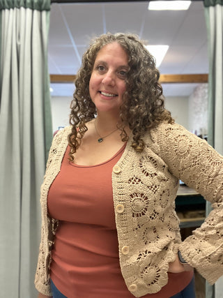 A smiling woman with curly brown hair wearing a beige crochet cardigan over a terracotta-colored tank top, standing in an indoor room with curtains and a mirror visible in the background.