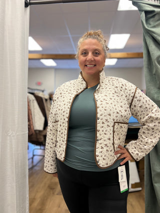 A woman wearing a cream-colored cropped jacket with a brown floral pattern and brown trim, layered over a sage green top and black leggings, standing in a fitting room.