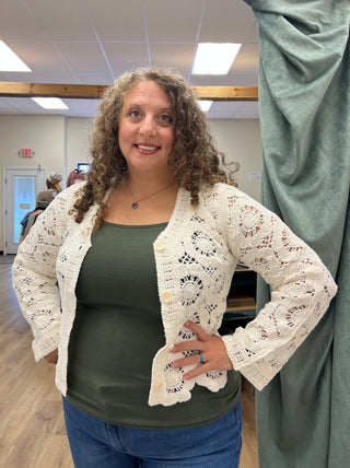 A woman with curly brown hair wearing a cream-colored, hand-crocheted lace cardigan over an olive green top and blue jeans, standing in an indoor room with a green curtain on the right.