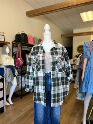 A green and white plaid button-up shirt displayed on a white mannequin torso, styled with a light pink top and blue jeans inside a clothing boutique.