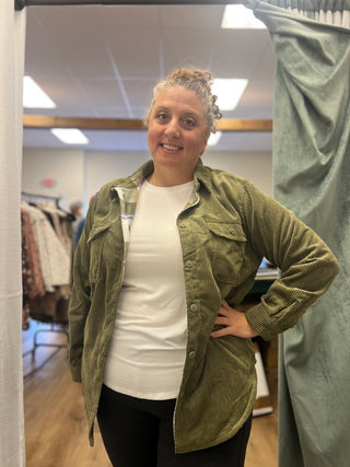 A woman posing in a fitting room wearing an olive green corduroy button-up shirt over a white t-shirt and black pants.