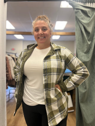 A woman wearing a green and white plaid flannel shirt over a white t-shirt and black pants, standing in a fitting room.