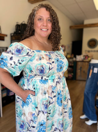 A woman wearing a white off-the-shoulder dress with a blue and teal floral print, standing in a retail store.