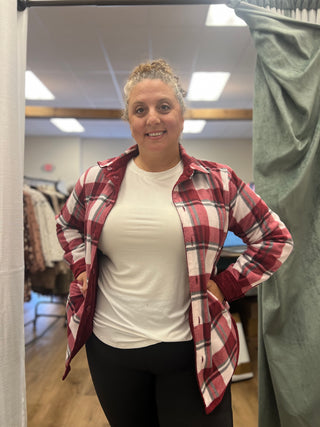 A woman posing in a fitting room wearing a red, white, and black plaid button-up shirt over a white t-shirt and black pants.