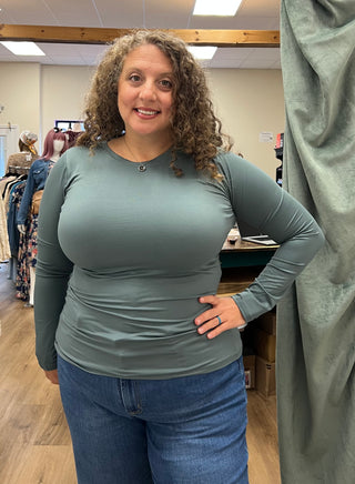 A woman with curly brown hair wearing a fitted sage green long-sleeve top and blue jeans, standing in a clothing store.