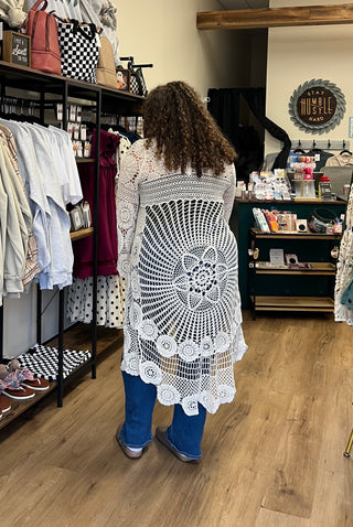 A person standing in a boutique, viewed from behind, wearing a white crochet cardigan with a large circular floral pattern over blue jeans.