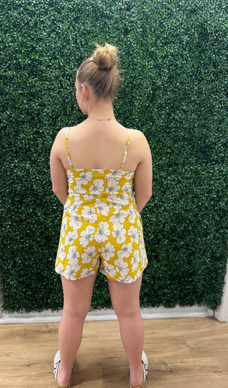 A woman stands with her back to the camera wearing a yellow and white floral romper in front of a green hedge wall.