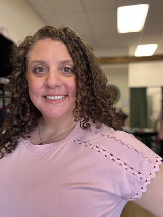 A smiling woman with curly brown hair wearing a light pink short-sleeve t-shirt with scalloped eyelet detailing on the shoulder, photographed in an indoor setting.