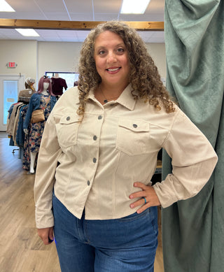 A woman with curly brown hair wearing a beige corduroy button-up shirt and blue jeans, standing in a clothing boutique with mannequins and a green curtain in the background.