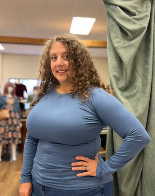A woman with curly brown hair wearing a fitted, long-sleeve dusty blue top and blue jeans, standing in a boutique setting with a green curtain on the right.