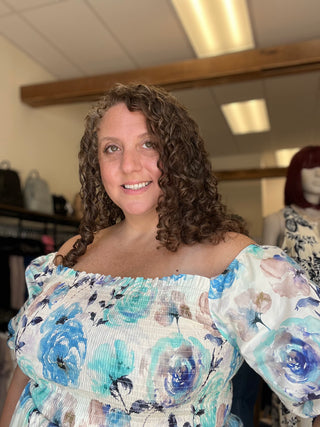 A smiling woman with curly brown hair wearing a blue and white floral off-the-shoulder top, standing in what appears to be a clothing boutique.