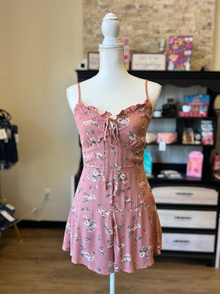 A pink floral romper displayed on a white mannequin torso in a retail store. The romper features thin spaghetti straps, a ruffled neckline, a front tie detail, and a flared skirt with an allover floral pattern.