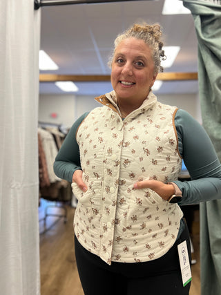 A woman wearing a cream quilted vest with a brown floral pattern over a sage green long-sleeve shirt, standing in a retail fitting room.
