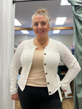 A woman wearing a cream-colored lace-trimmed cardigan over a beige top and black leggings, standing in a fitting room or boutique.