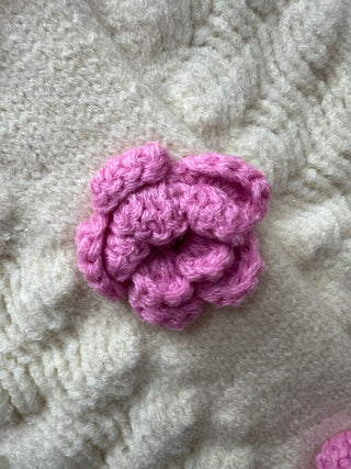 A close-up of a pink crocheted flower applique attached to a chunky cream-colored knit fabric.