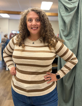 A woman with curly brown hair wearing a beige and brown horizontally striped long-sleeve knit top and blue jeans, standing in a fitting room with a green curtain in the background.