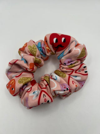 A pink fabric scrunchie featuring a playful cartoon pattern with red, orange, blue, and purple characters, accented with gold glitter patches and topped with a red plush heart character.
