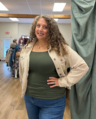 A woman with curly brown hair stands in a boutique wearing a beige corduroy jacket over an olive green tank top and blue jeans. She is posing in front of a green curtain inside the store.