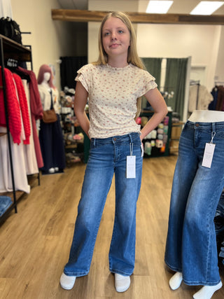 A young woman stands in a clothing boutique wearing blue wide-leg jeans and a cream floral short-sleeve top. A mannequin to her right displays another pair of blue wide-leg jeans with a price tag attached.
