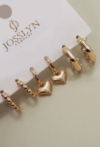 Six Josslyn Company gold-toned earrings displayed on a white branded card, including hoop earrings and heart-shaped charms, photographed on a light neutral surface.