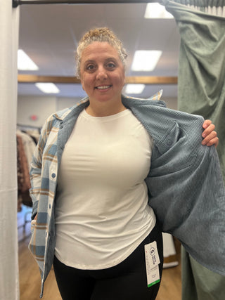 A woman in a fitting room wearing a white crewneck t-shirt and black leggings, holding open a blue corduroy button-up shirt over a blue and white plaid shirt.