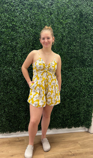A young woman stands in front of a green artificial hedge wall wearing a yellow and white floral romper with white sneakers.