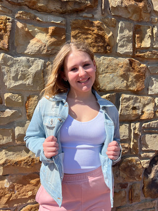A young woman lying on a stone wall, wearing a light blue denim jacket over a white ribbed tank top and pink pants.