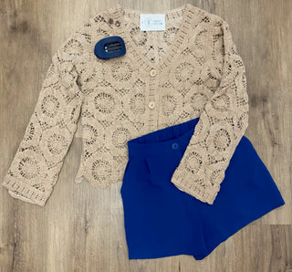 A beige crochet cardigan sweater with a lace pattern and wooden buttons, paired with royal blue shorts, laid flat on a wood floor. A small blue hair clip is also placed on the cardigan.