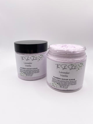 Two jars of The She Shall Lavender Vanilla Foaming Sugar Scrub on a white background, with one jar closed in black packaging and the other open showing the light purple scrub inside.