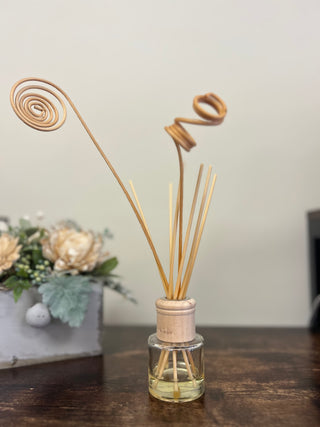 A reed diffuser bottle with a small clear glass container, light wood stopper, and several natural reed sticks, including two spiral-shaped reeds, displayed on a dark wooden surface with a blurred floral arrangement in the background.