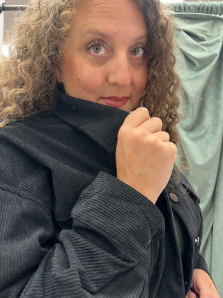 A close-up shot of a woman with curly light brown hair wearing a dark gray ribbed corduroy jacket, posing in front of a sage green curtain.