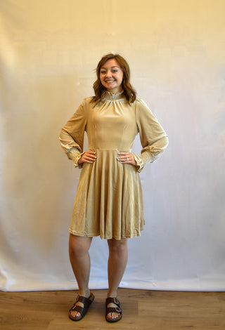 A woman modeling a beige long-sleeve mini dress with a gathered waist and flared skirt, paired with black slide sandals, standing against a white studio backdrop.