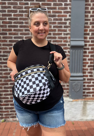 A woman wearing a black t-shirt and denim shorts models a black and white checkered fanny pack with gold-tone hardware, standing in front of a brick wall.