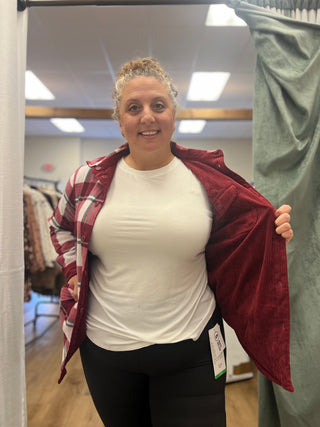 A woman in a fitting room wearing a white t-shirt and black leggings, holding open a red and white plaid flannel shirt to show a burgundy corduroy-lined jacket inside.