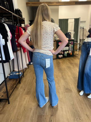A woman with long blonde hair stands with her back to the camera wearing light blue flare jeans and a cream floral t-shirt inside a clothing boutique.