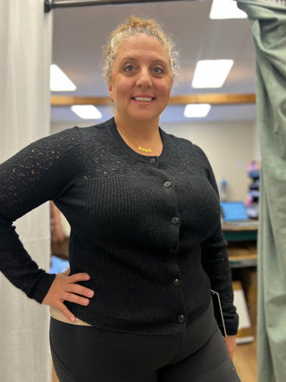 A woman wearing a black long-sleeve button-up cardigan with lace detailing on the upper chest and sleeves, paired with black leggings, standing in an indoor room with a mirror and clothing setup visible in the background.