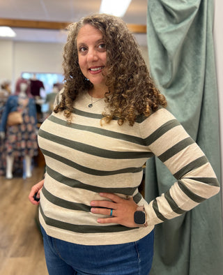 A woman with curly brown hair wearing a beige and dark green striped long-sleeve knit top, standing in a boutique with a green curtain behind her.