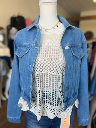 A mannequin wearing a light blue denim jacket over a white crochet lace top, paired with blue jeans and a gold necklace, in what appears to be a clothing store.