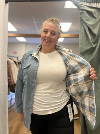 A woman in a fitting room wearing a light blue corduroy shirt jacket over a white t-shirt and black pants, holding open a light blue, white, and tan plaid shirt to show the lining.