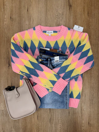 A pink, yellow, and blue chevron-patterned knit sweater is laid flat on a wood floor, styled with folded blue jeans, a beige handbag, and a white tag.
