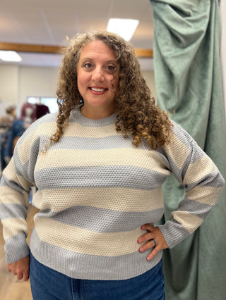 A woman with curly brown hair wearing a cream and light blue-gray striped waffle-knit sweater and blue jeans, standing in a clothing store.