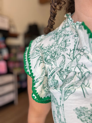 Close-up of a person wearing a white short-sleeve top with green toile-style botanical and bird illustrations and green scalloped trim.